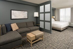 A hotel room with a gray sofa, a small wooden bench, a bed with white bedding, a nightstand with a lamp, and a large window with curtains. - Hyatt Place Bentonville/Rogers (Rogers)