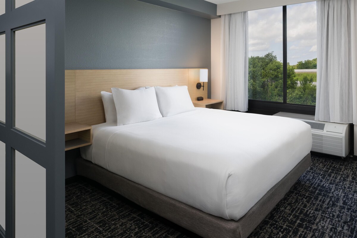 Une chambre d’hôtel avec un grand lit, une tête de lit, une table de chevet, une commode, une fenêtre avec des rideaux et un appareil de climatisation.