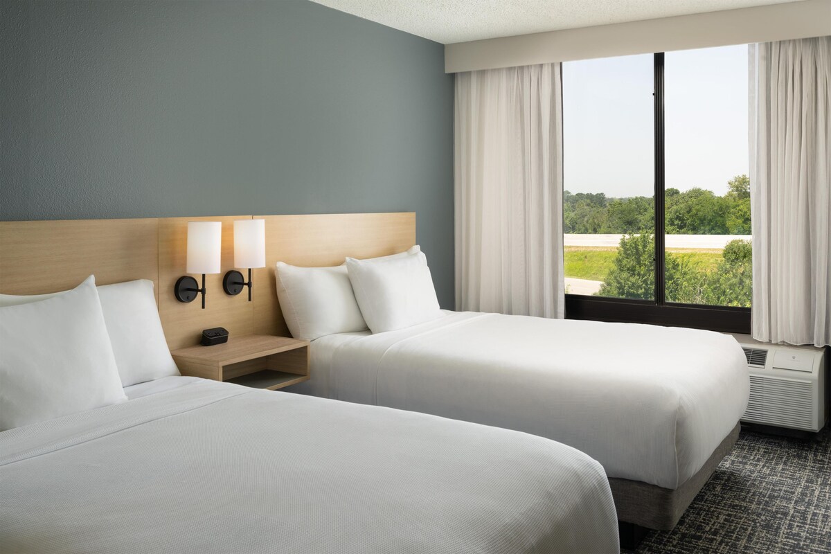 Une chambre d’hôtel avec deux lits, une table de chevet, une lampe fixée au mur et une fenêtre avec des rideaux.