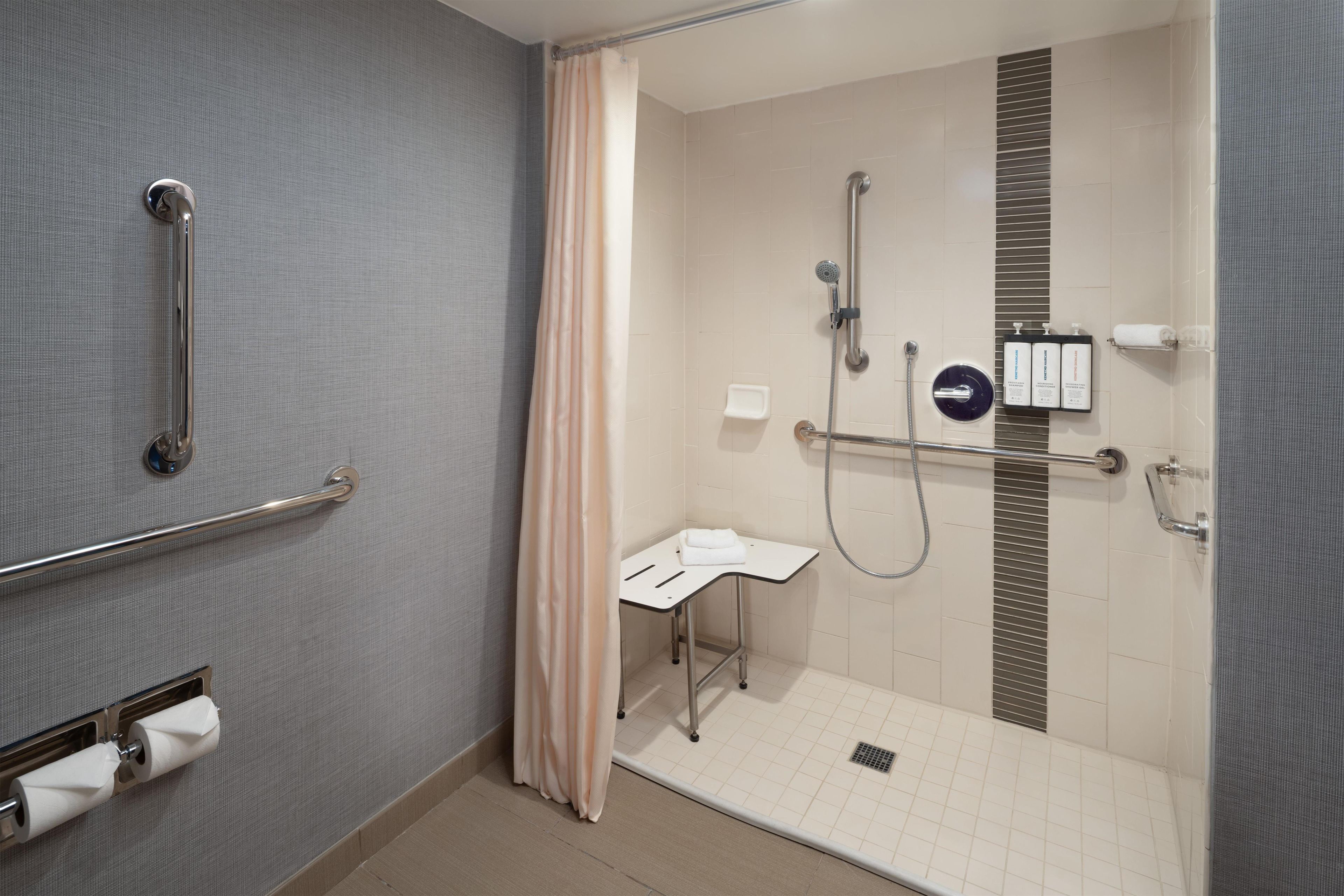 Ein modernes Badezimmer mit bodengleicher Dusche, Haltegriffen, einer Toilette mit Handlauf und einem Waschbecken mit Handtuchhalter.