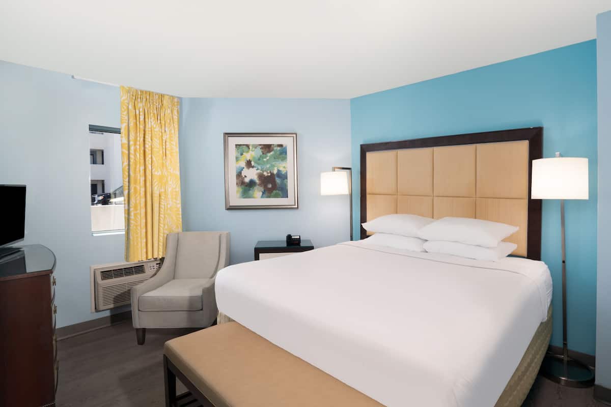 A hotel room with a large bed, a chair, a desk, a TV, and a painting on the wall.