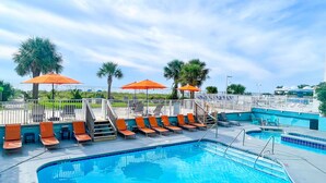 A swimming pool with sun loungers and orange umbrellas, surrounded by palm trees and a clear sky. - Palette Resort Myrtle Beach (Myrtle Beach)