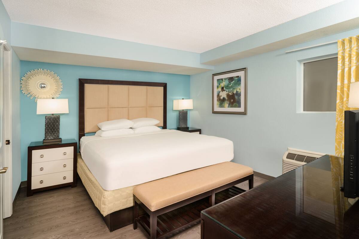 A hotel room with a large bed, two bedside tables with lamps, a bench, a TV, and a window with curtains.
