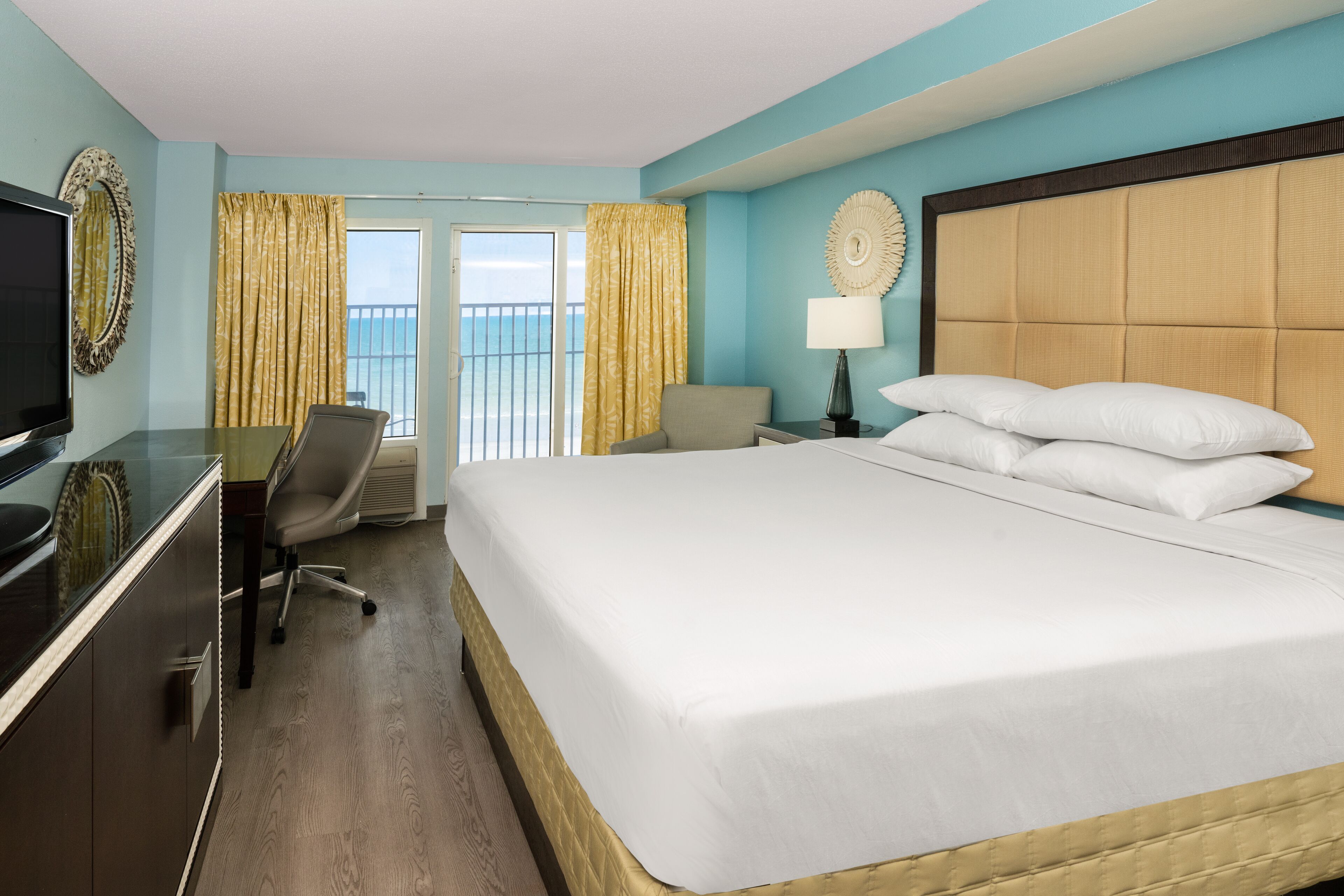 Habitación de hotel con una cama grande, un escritorio con televisor, una silla, una lámpara y vistas al exterior mediante una puerta corredera.