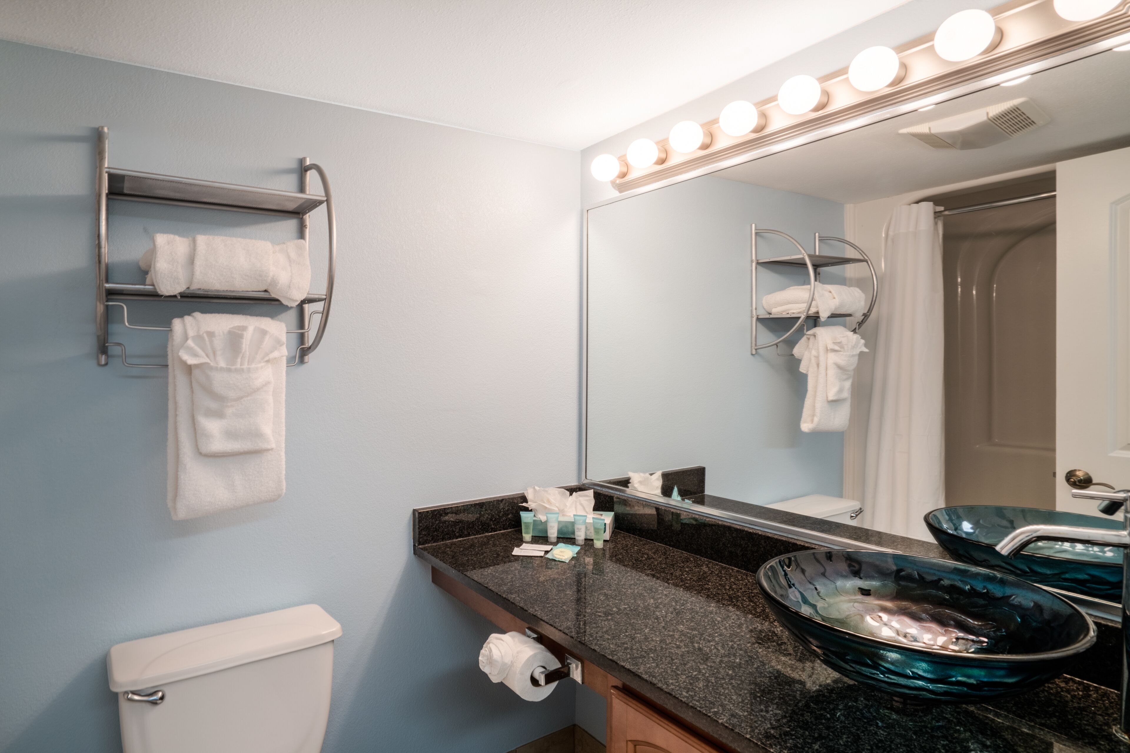 A bathroom with a toilet, a sink with a blue glass basin, a towel rack, and a mirror with lights.