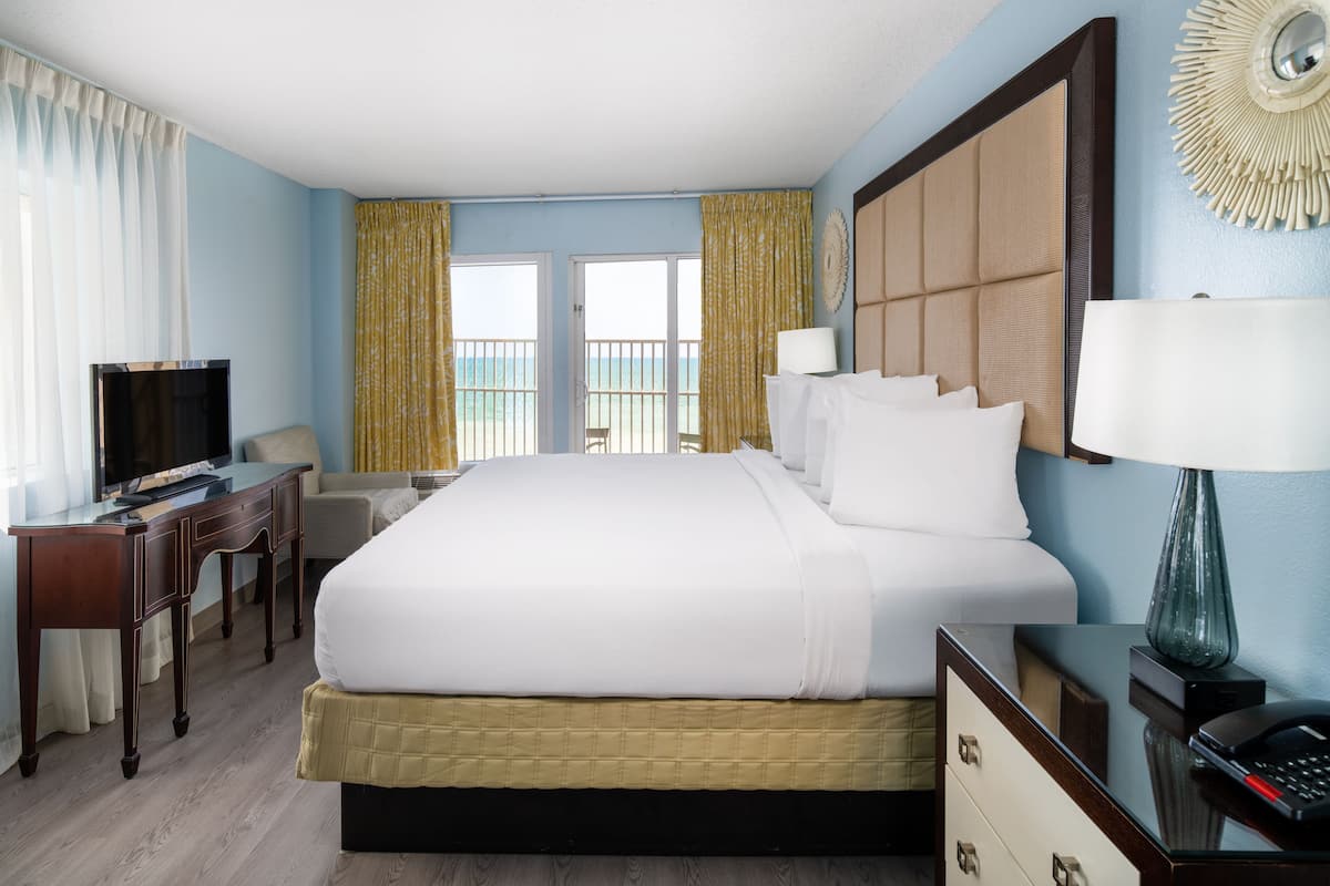 A hotel room with a large bed, a desk with a TV, a chair, and a view of the ocean through the windows.