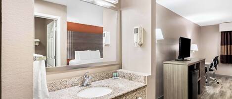 A hotel bathroom with a granite sink, a mirror, a towel rack, a hairdryer, and a television on a desk.