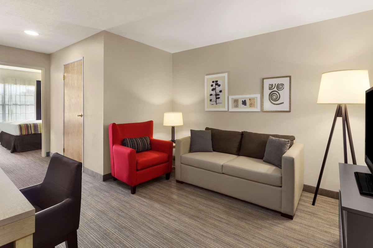 A hotel room with a red armchair, a beige sofa, a desk with a lamp, and a TV stand.