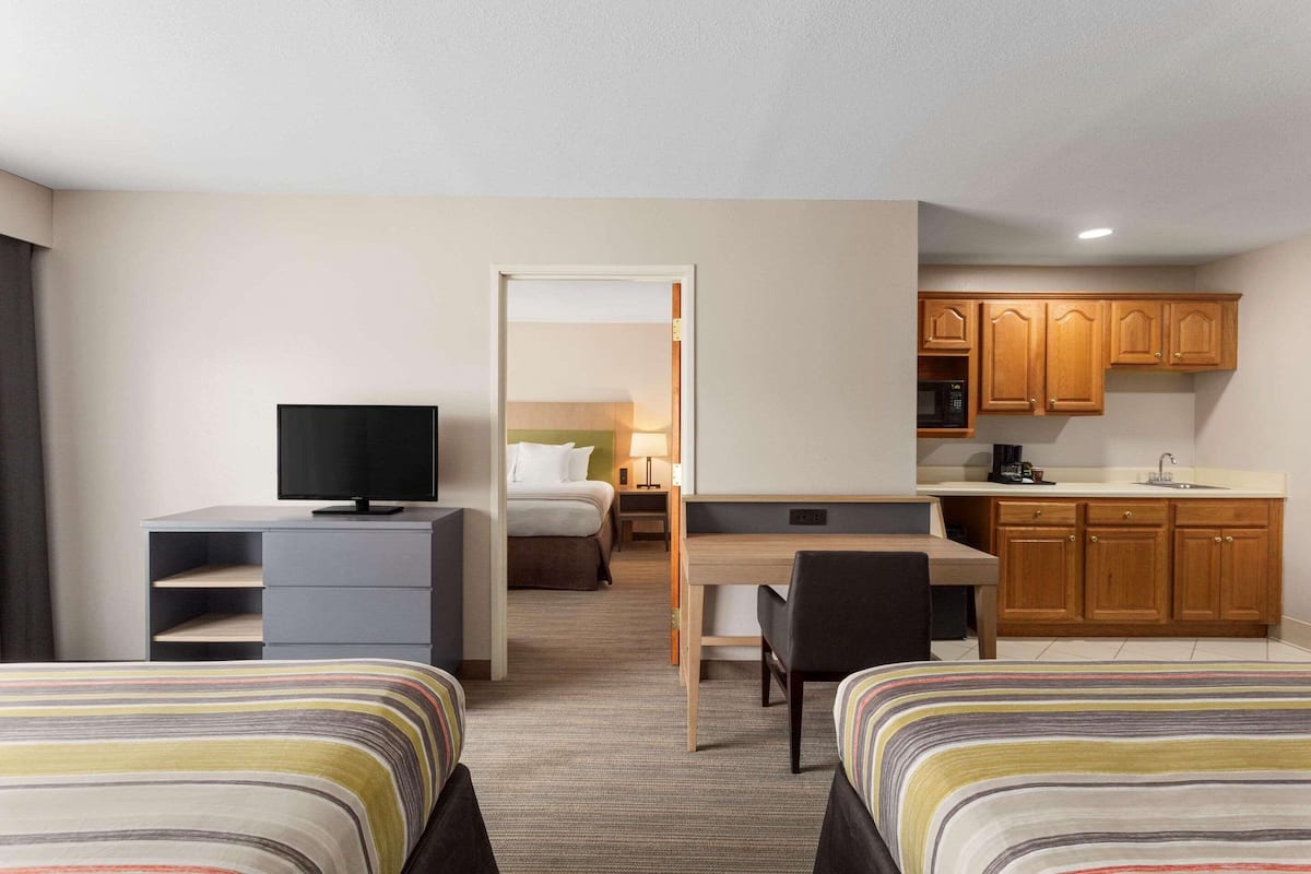 A hotel room with two beds, a desk, a television, a kitchenette, and a bathroom visible through an open door.
