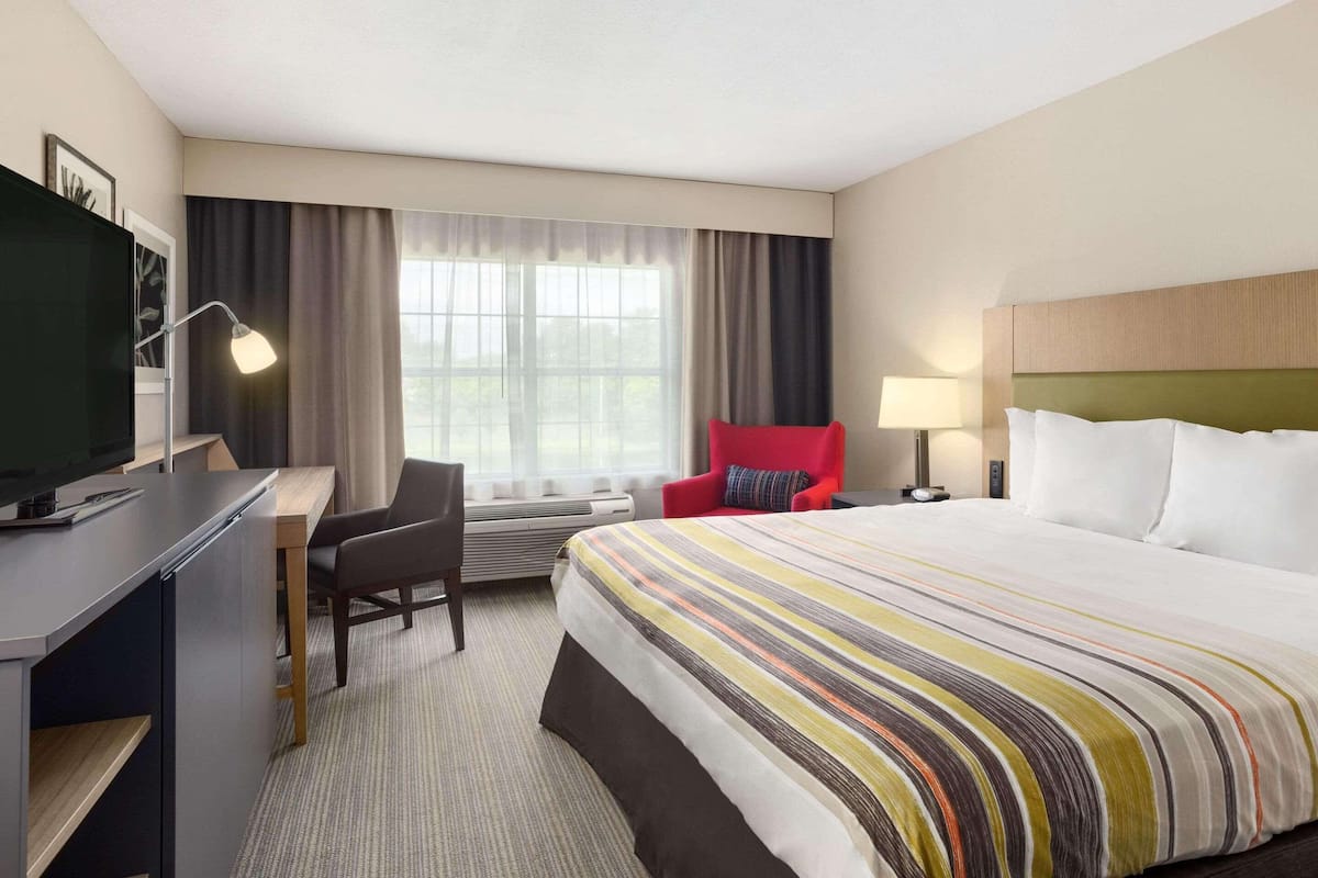 A hotel room with a bed, a desk with a computer, a chair, a TV, a window with curtains, and a lamp.