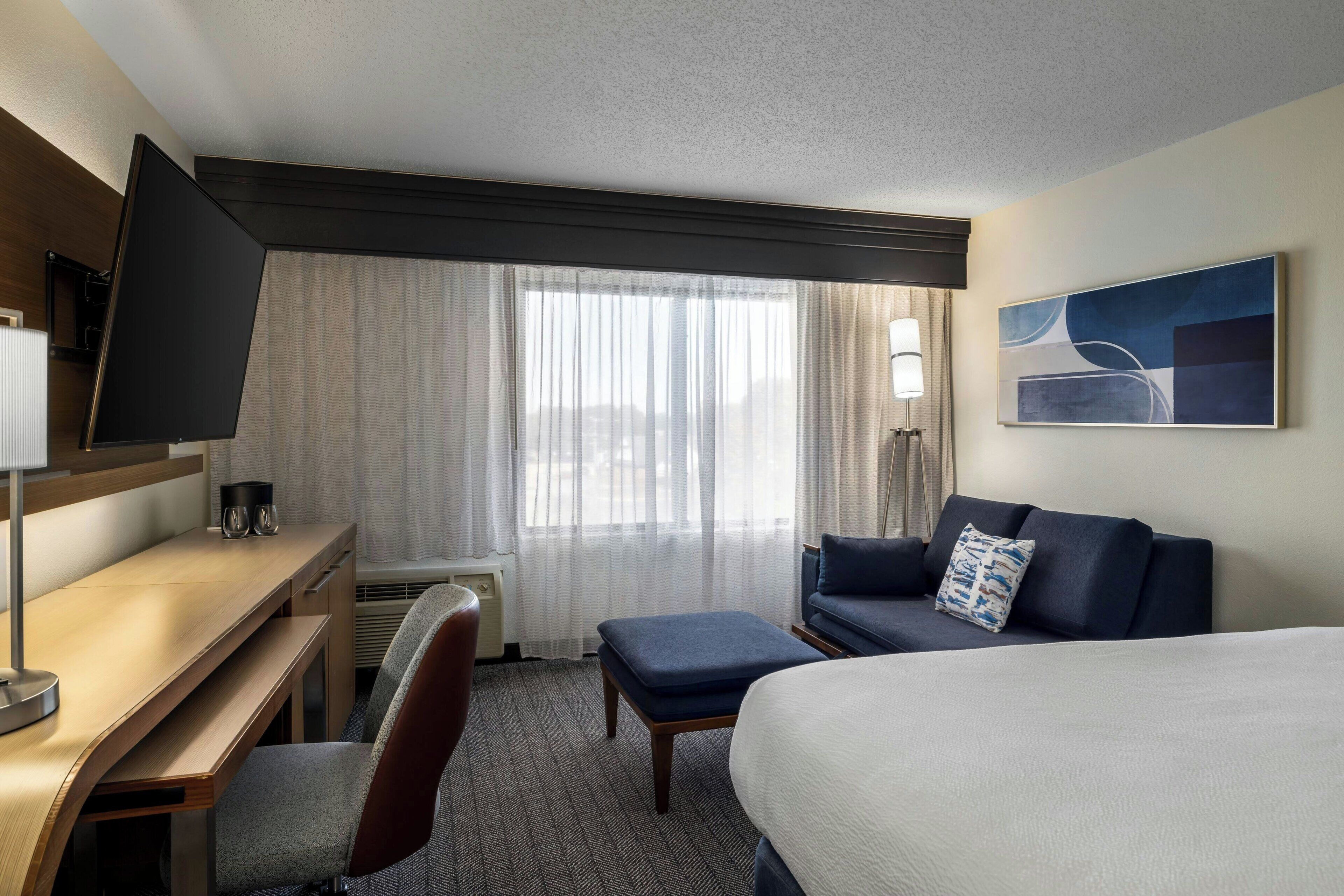 A hotel room with a bed, a desk, a chair, a sofa, a TV, and a window with curtains.