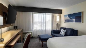 A hotel room with a bed, a desk, a chair, a sofa, a TV, and a window with curtains.