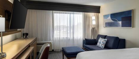 Quarto de hotel com cama, escrivaninha, cadeira, sofá, televisão e janela com cortinas.