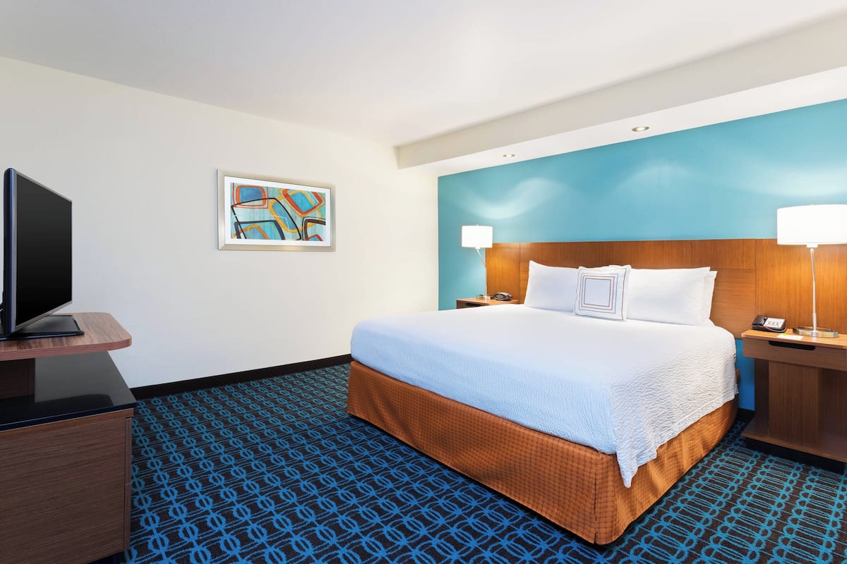 A hotel room with a large bed, a flat-screen TV, a painting on the wall, and a blue patterned carpet.