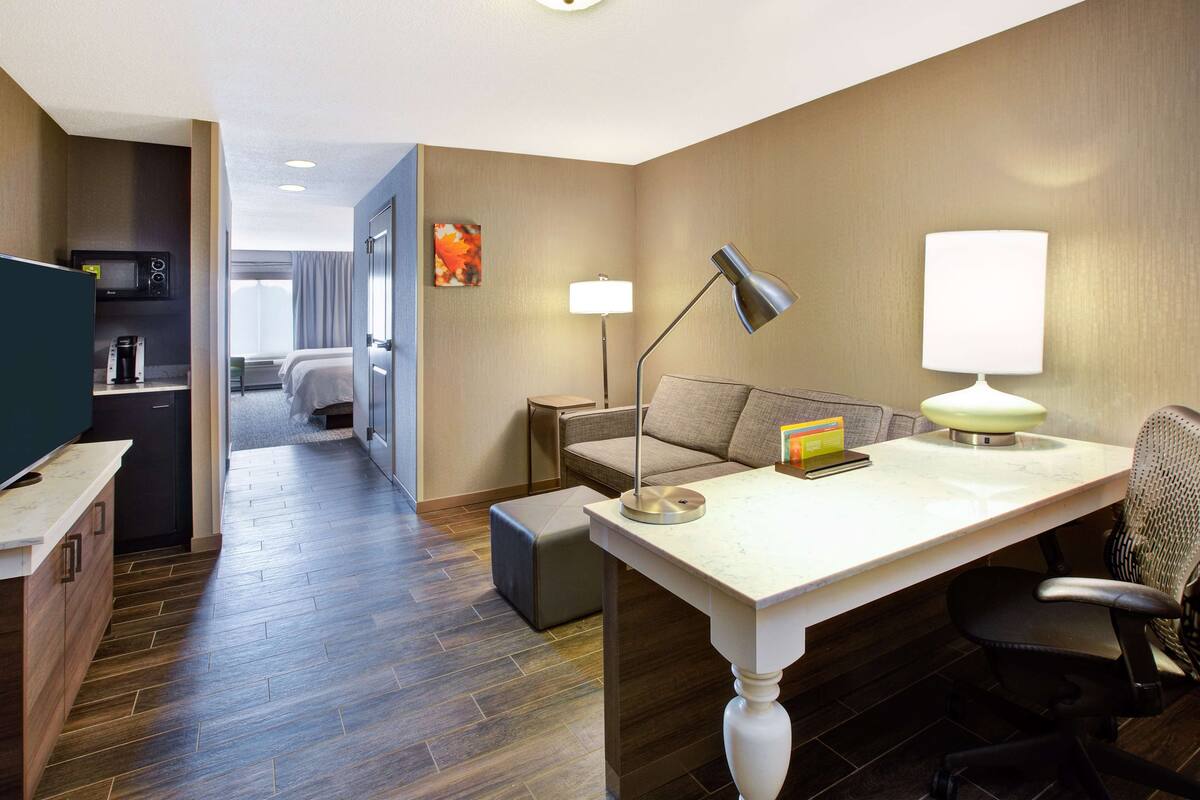 A hotel room with a kitchenette, a desk with a lamp, a chair, a bed, and a view of the hallway.