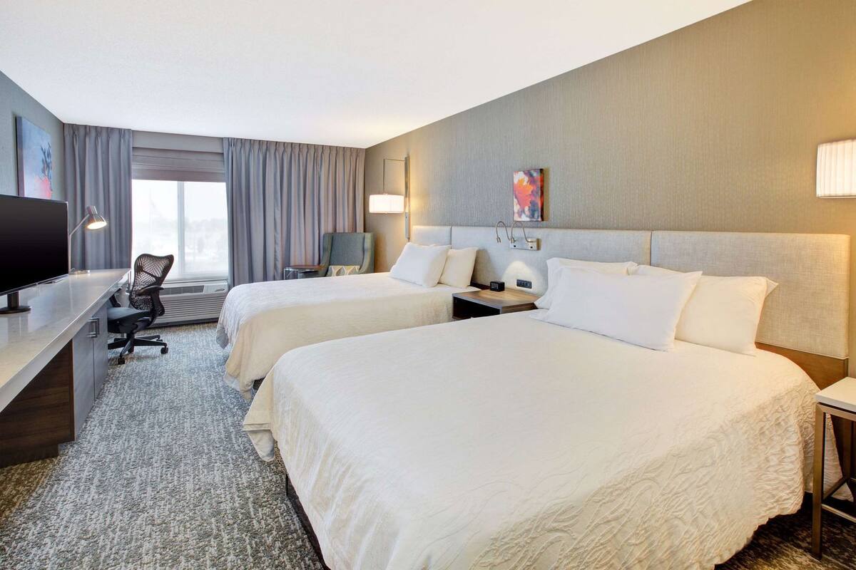 A hotel room with two beds, a desk, a chair, a TV, and a large window.