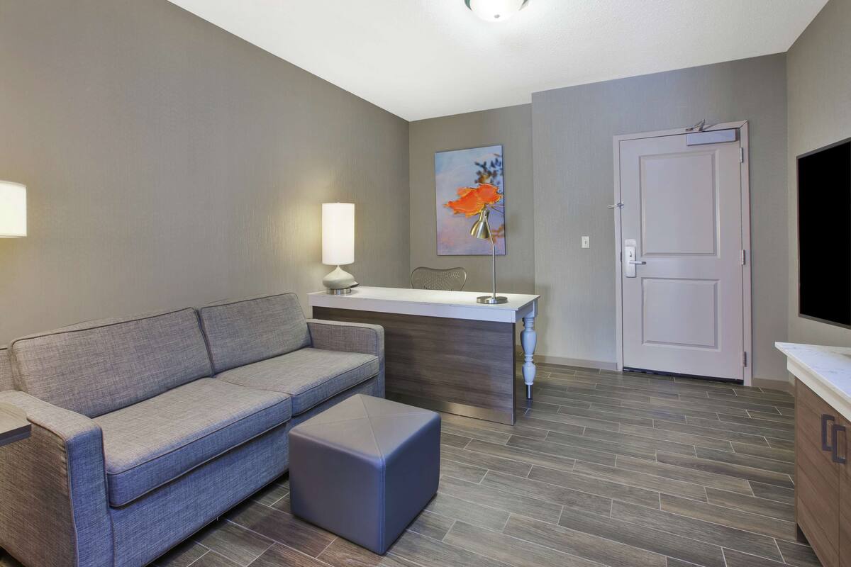 A modern living room with a grey sofa, a small ottoman, a white desk, and a door.