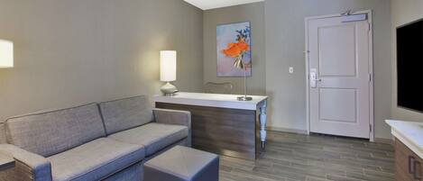 A modern living room with a grey sofa, a small ottoman, a white desk, and a door.