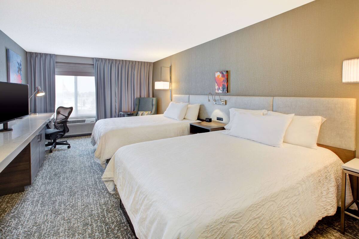 A hotel room with two beds, a desk with a computer, a chair, a TV, and a window with curtains.