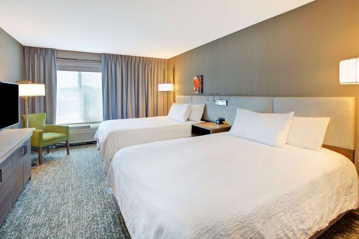 A hotel room with two beds, a desk, a chair, a TV, and a window with curtains.