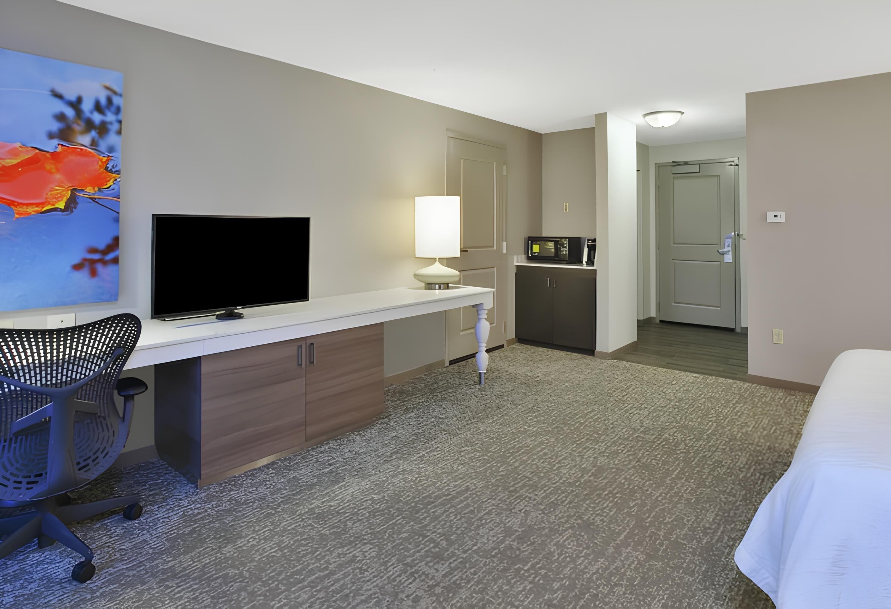 A hotel room with a desk, TV, lamp, kitchenette, and a bed.