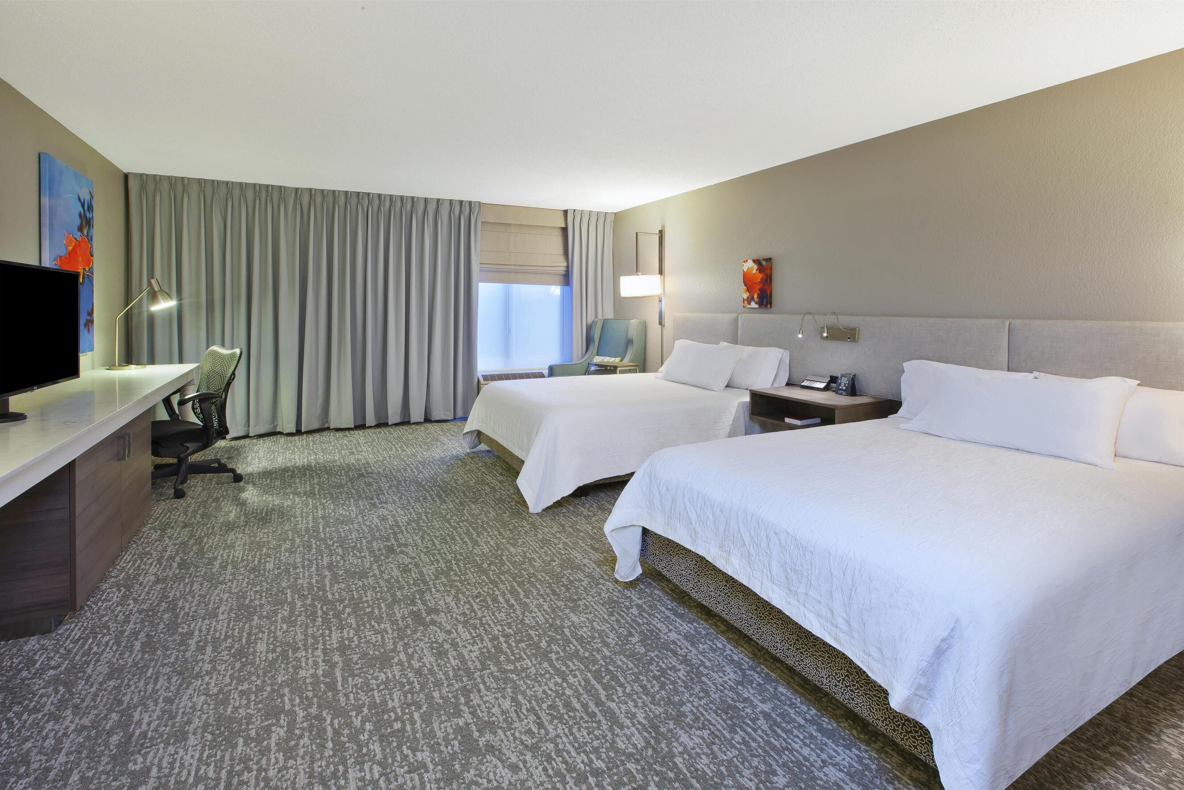 A hotel room with two beds, a desk with a TV, a chair, and a window with curtains.