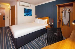 A hotel room with a bed, a desk, a chair, a hanging coat rack, and a painting on the wall.