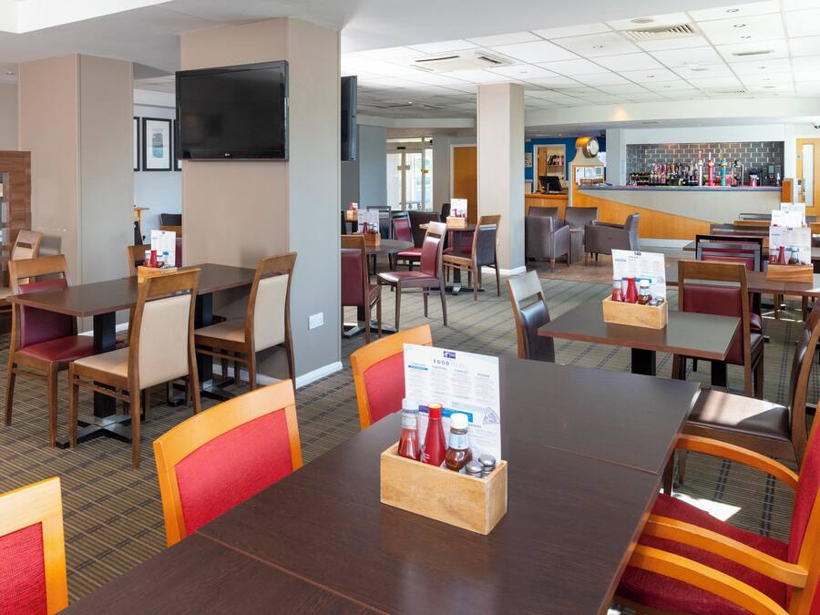 Uma área de refeições de hotel com várias mesas e cadeiras, um bar e uma televisão de tela plana na parede.