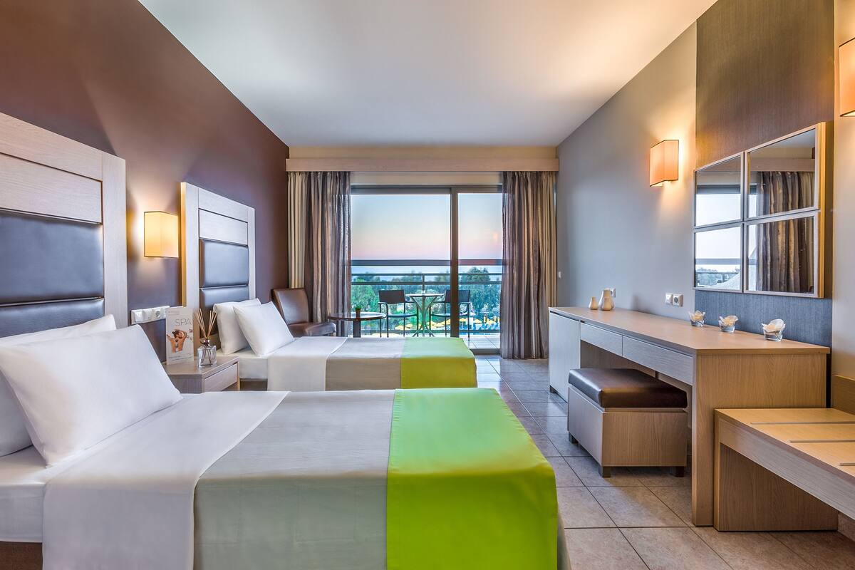 Habitación de hotel con dos camas, un escritorio, una silla, una mesita y un balcón con vistas.