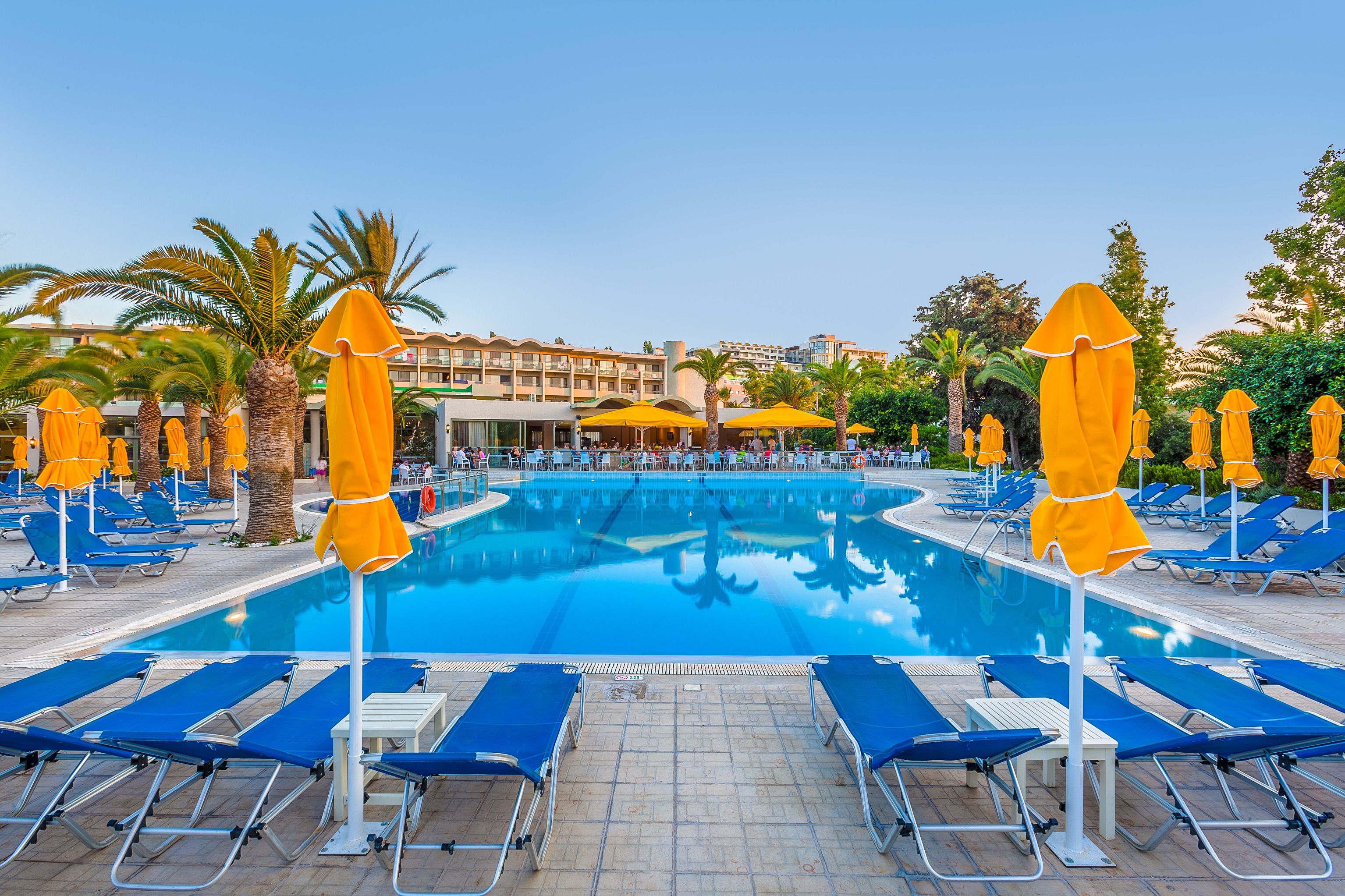 Une piscine entourée de palmiers et, au loin, d’un bâtiment d’hôtel, avec des chaises longues bleues et des parasols jaunes.