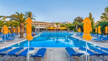 Une piscine entourée de palmiers et, au loin, d’un bâtiment d’hôtel, avec des chaises longues bleues et des parasols jaunes.