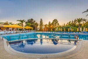 Una amplia piscina al aire libre con solárium de azulejos, rodeada de palmeras y sillones de descanso.