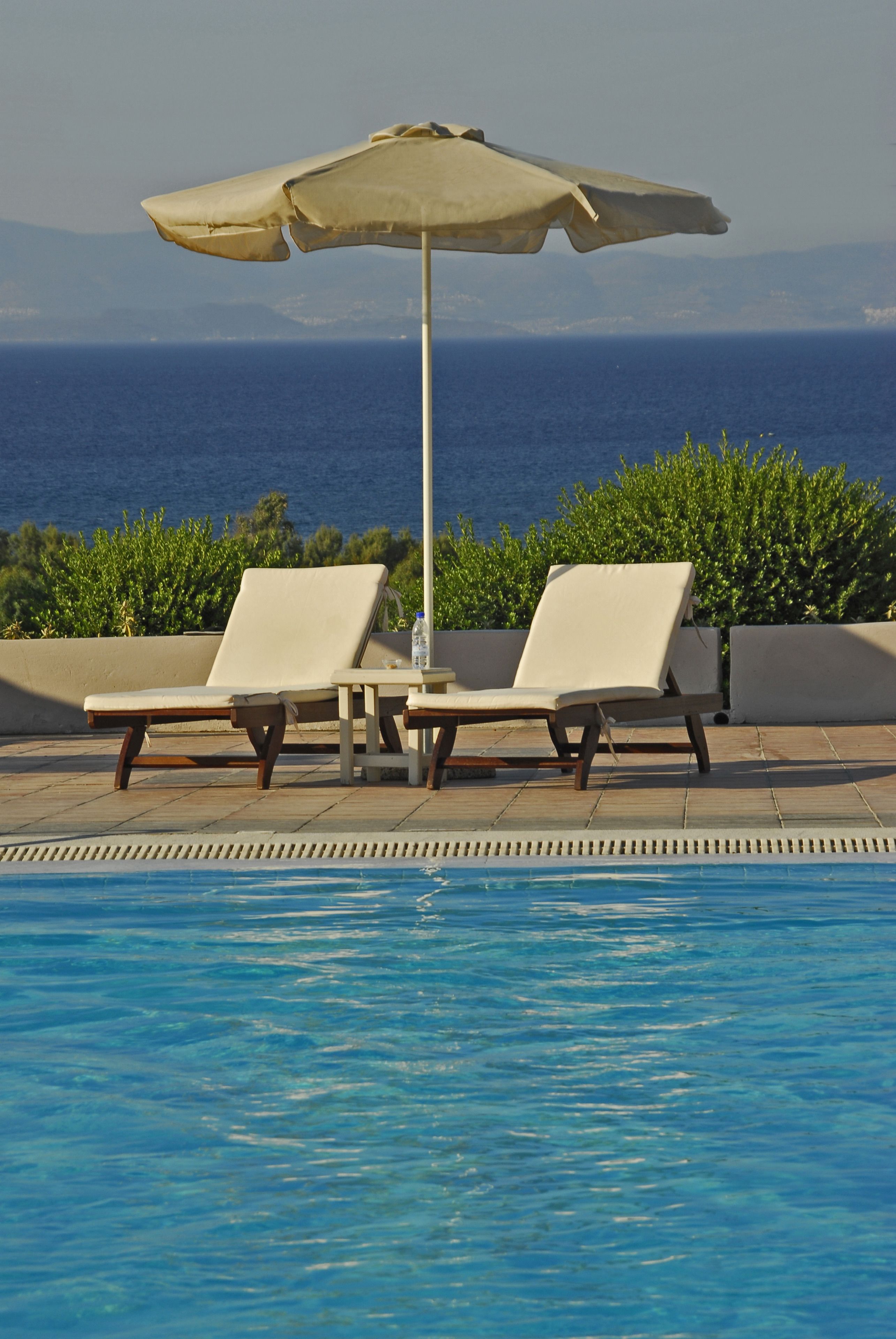 A poolside area with two lounge chairs and a large umbrella, overlooking a body of water.