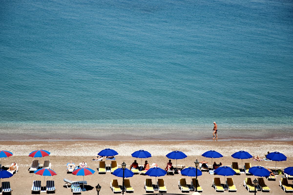 Une plage bordée de rangées de transats et de parasols, une mer d’un bleu limpide, et quelques personnes qui se promènent.