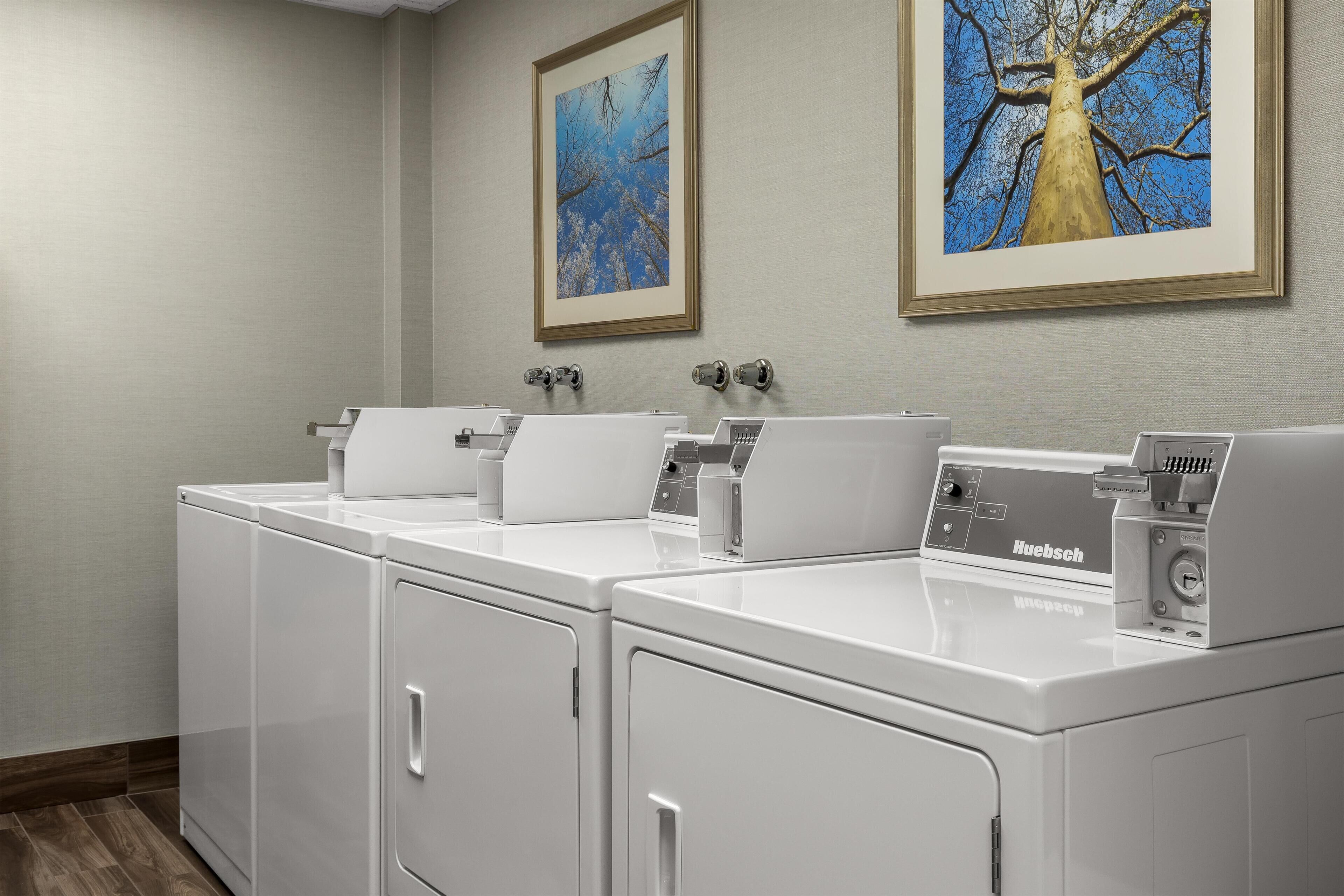 A laundry room with two white front-loading washers and dryers, a control panel, and two framed artworks on the wall.