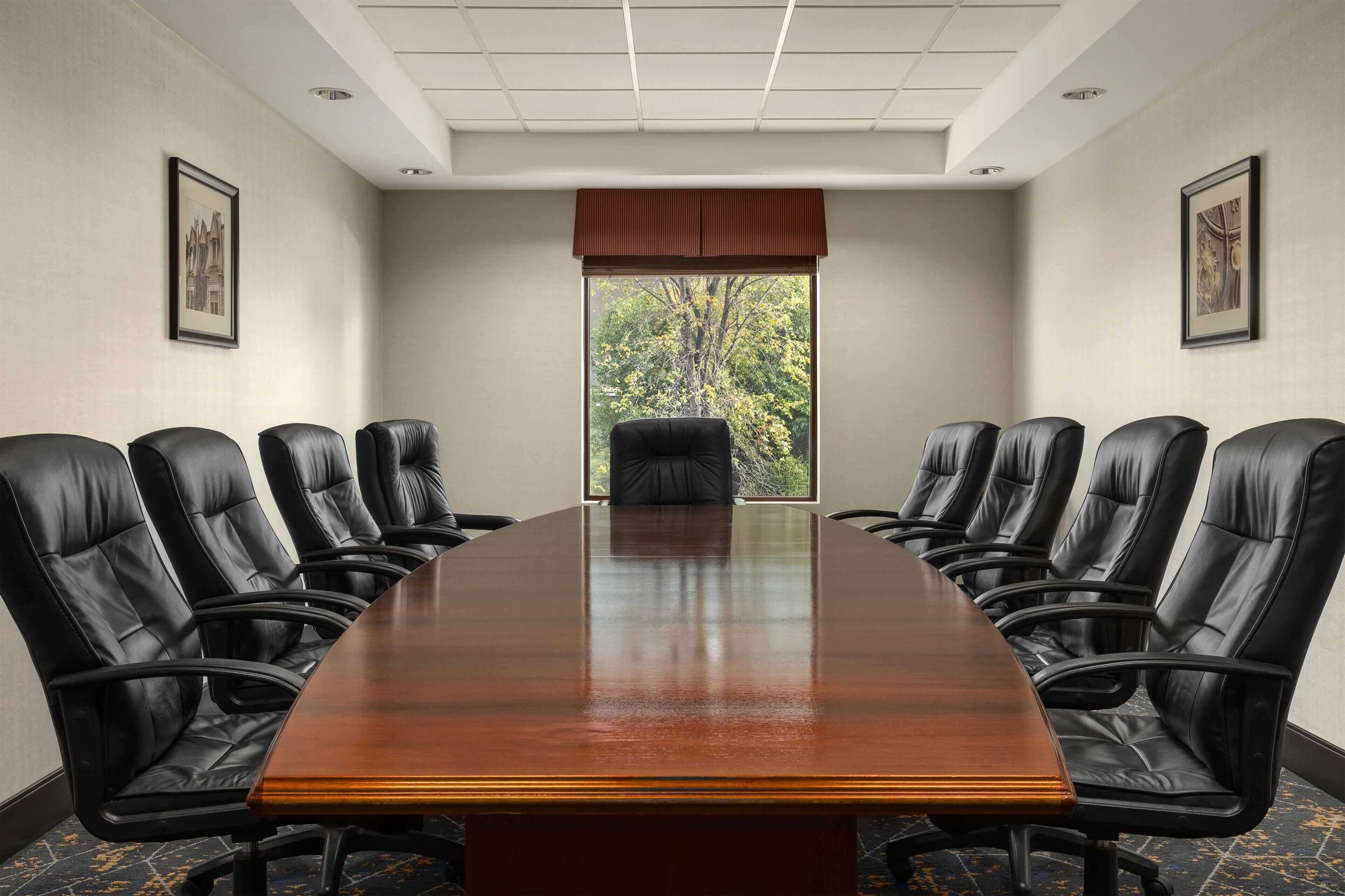 A conference room with a long table and black leather chairs, framed pictures on the walls, and a window with a view of trees.