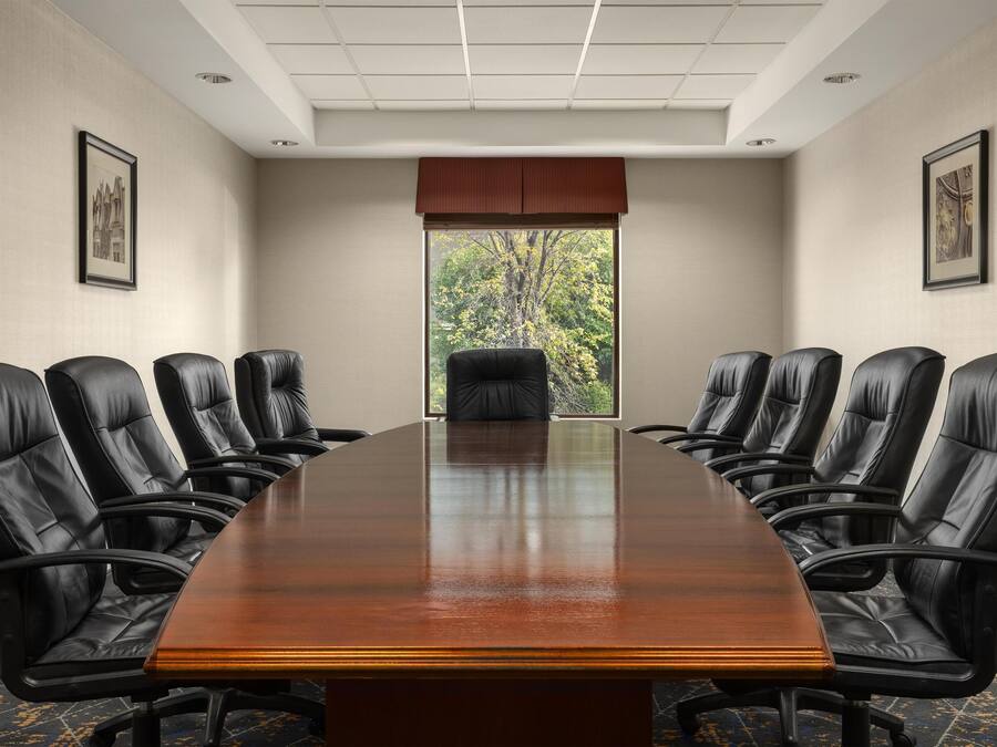 Un salón de conferencias con una larga mesa y sillas de cuero negro, cuadros en las paredes y una ventana con vista a árboles.