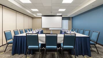 Un salón de conferencias con una larga mesa, sillas azules y una pantalla para proyector.
