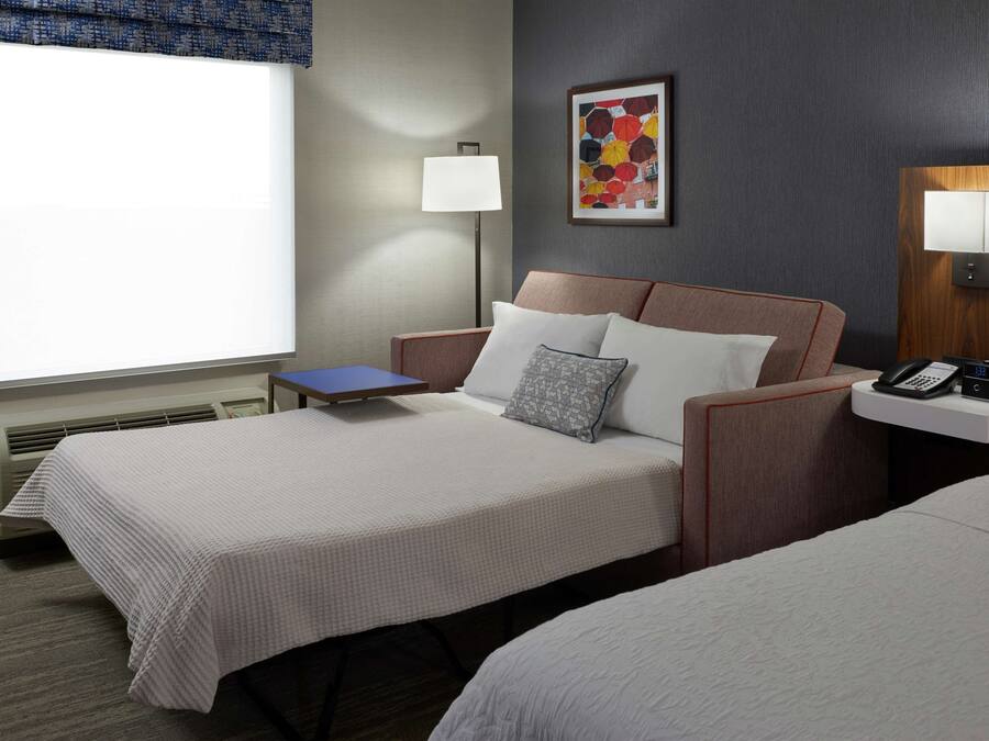 A hotel room with two beds, a bedside table with a lamp, a framed artwork on the wall, and a window with a view.
