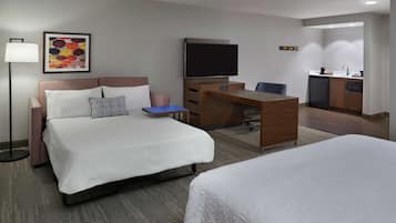 Quarto de hotel com duas camas, uma mesa de escritório, uma TV de tela plana, uma kitchenette e um abajur.