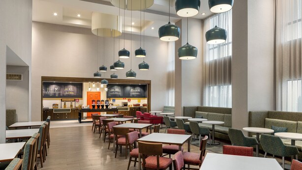 A modern hotel lobby with multiple seating areas, including tables and sofas, and a central food service area. - Hampton Inn & Suites by Hilton Montreal-Dorval (Dorval)