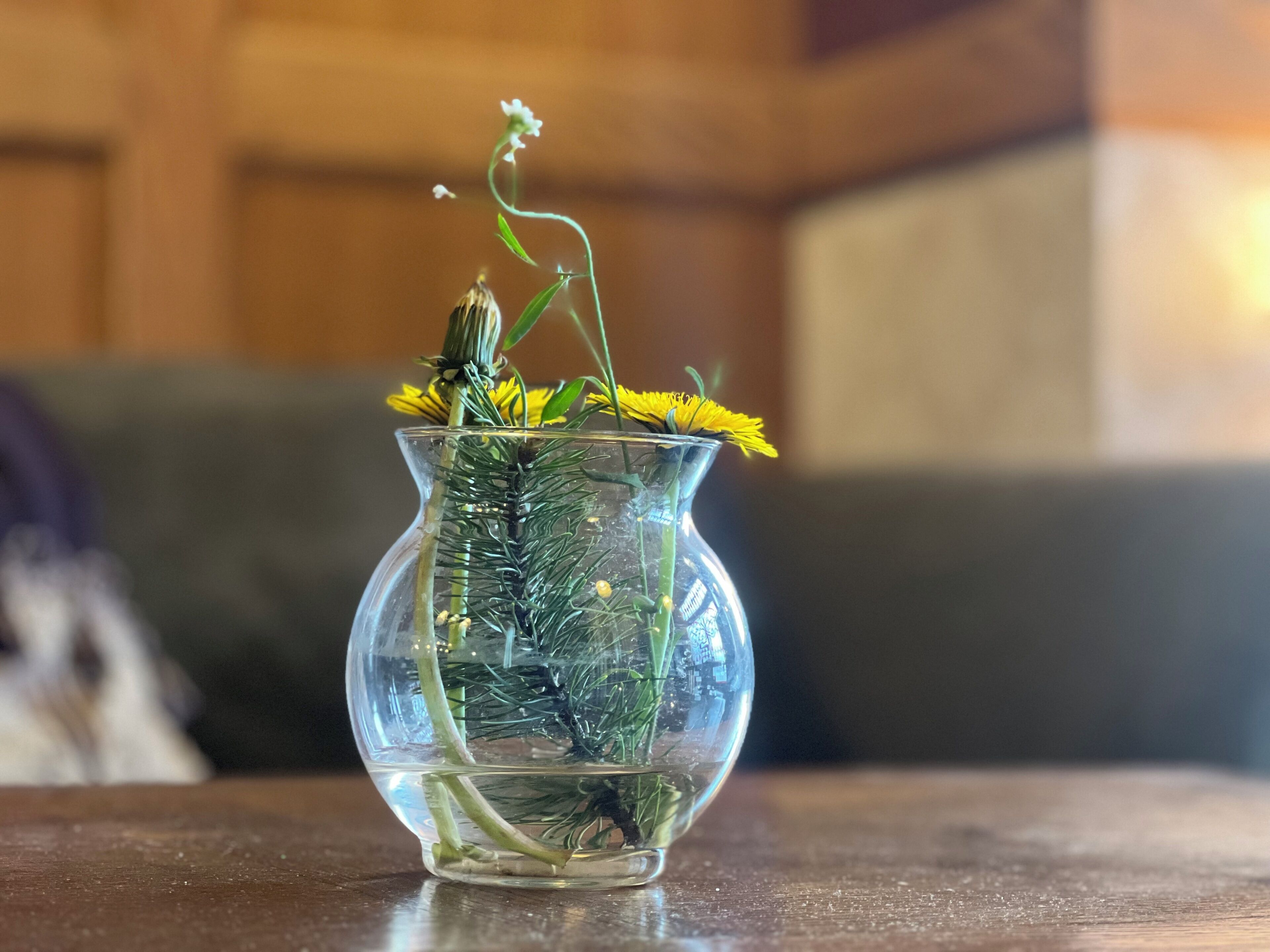 Un florero de vidrio transparente con follaje verde y flores amarillas sobre una superficie de madera.