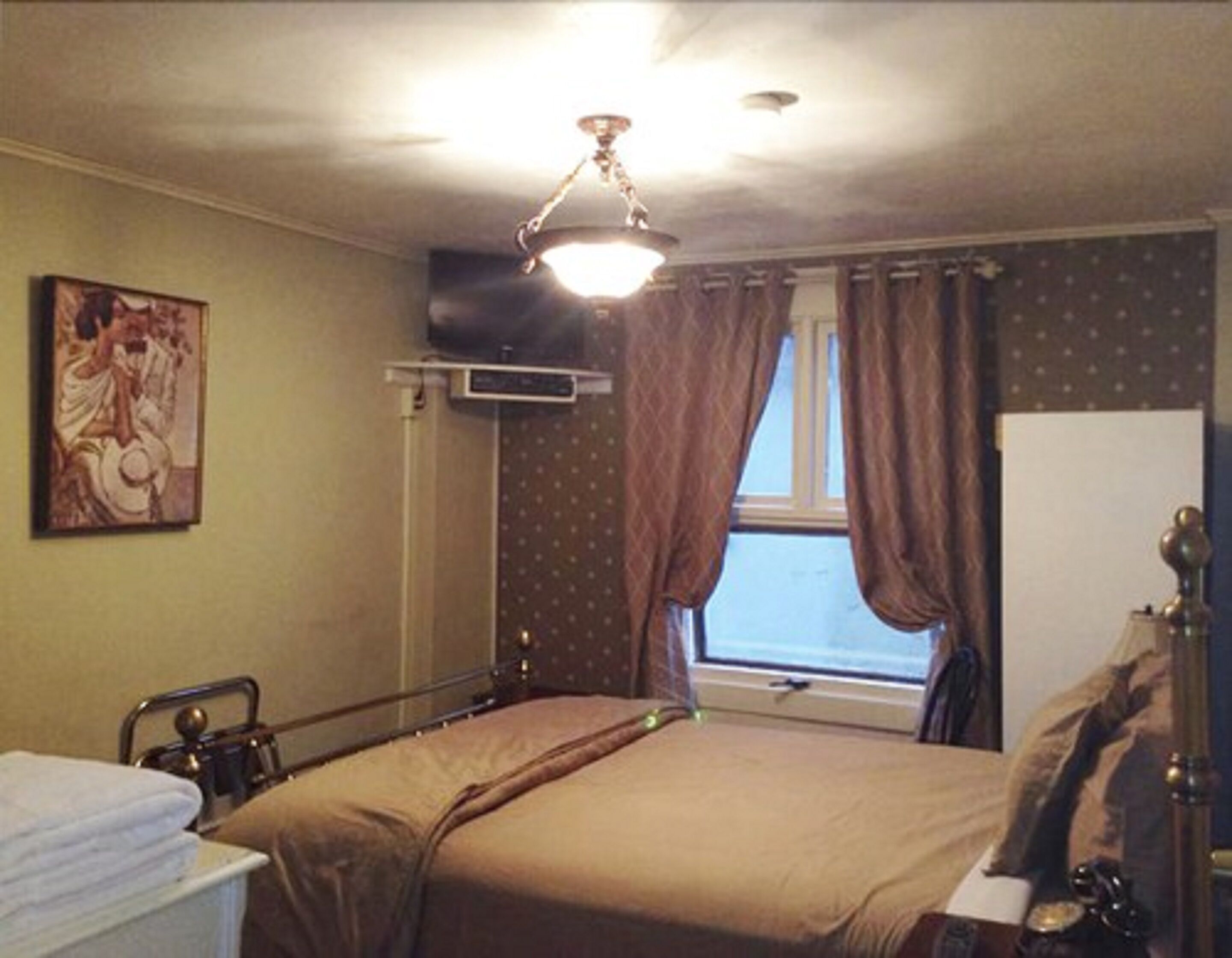 Una camera da letto con un letto, un comodino, una lampada, una finestra con tende, un quadro appeso al muro e una lampada a sospensione.