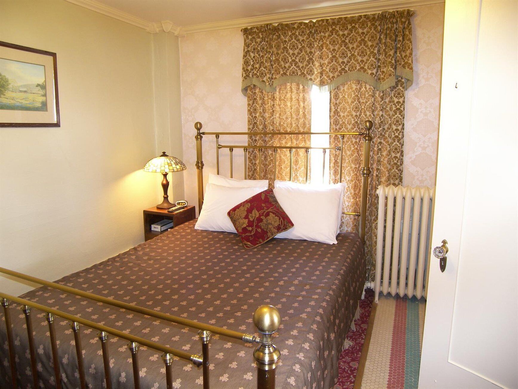 A bedroom with a bed, a nightstand, a lamp, a radiator, and a window with curtains.