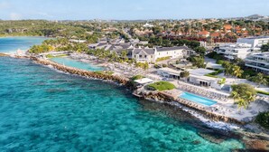 A coastal resort with a clear turquoise sea, a rocky shoreline, and a variety of buildings including a large white house with a pool. - Papagayo Beach Resort (Jan Thiel)