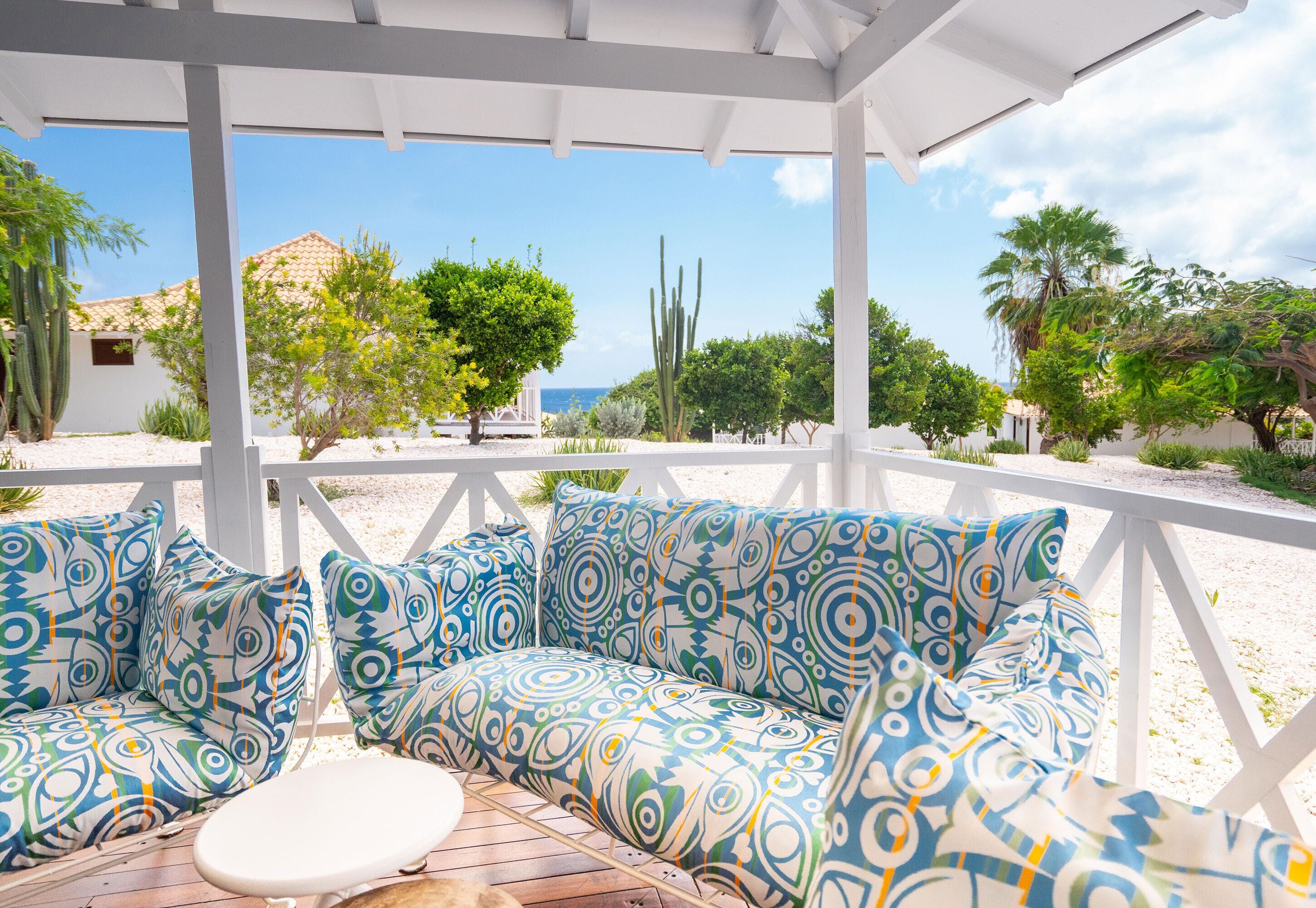 A covered outdoor seating area with patterned cushions and a wooden table, overlooking a garden with cacti and trees.
