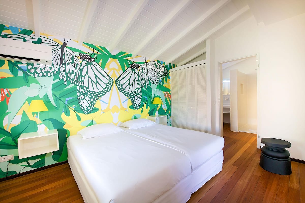 Habitación de hotel con cama, un mural en la pared, ventilador de techo y puerta que da a otra habitación.
