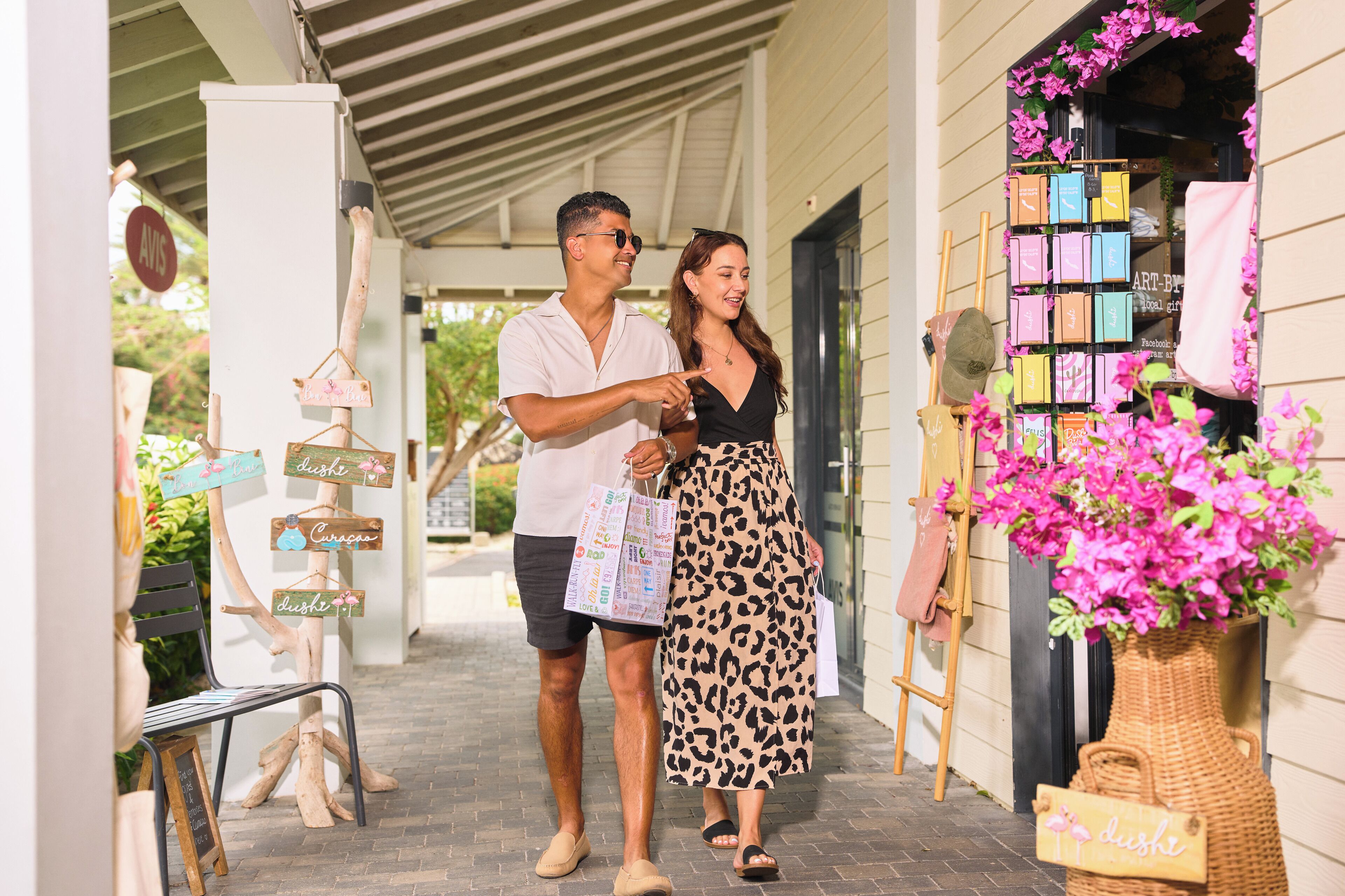 A couple walking past a boutique with a signboard, a display of colorful boxes, and a wicker basket with flowers.