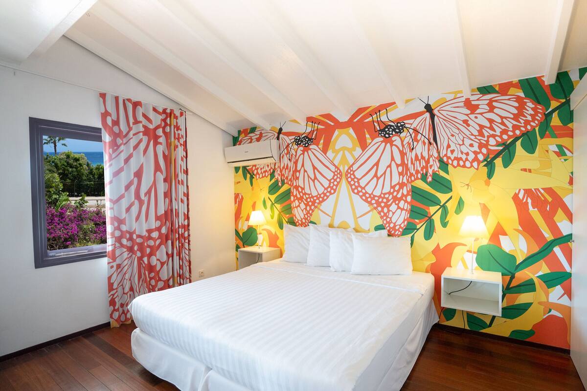 Un dormitorio con cama, ventana con cortinas, pared con diseño de hojas y mesita de noche con lámpara.