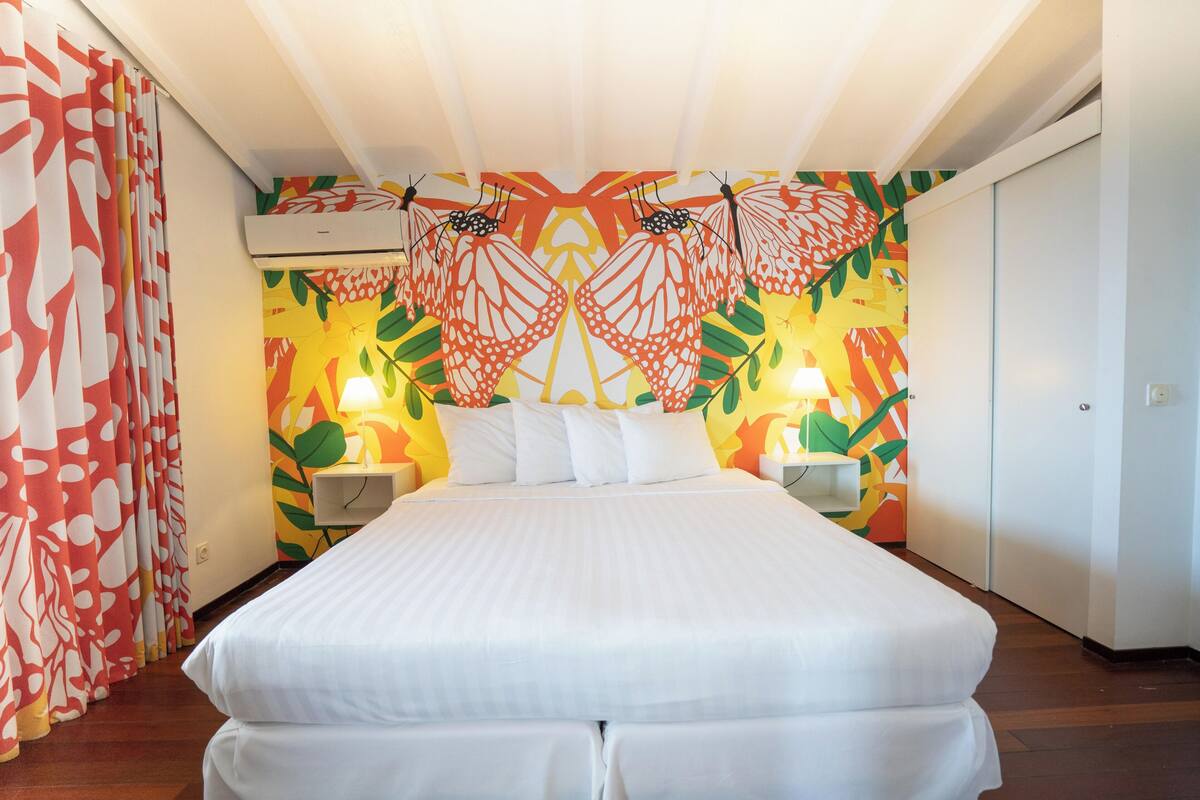 Habitación de hotel con una cama grande, dos mesitas de noche con lámparas, una pared con un mural de mariposas de colores y un ventilador de techo.